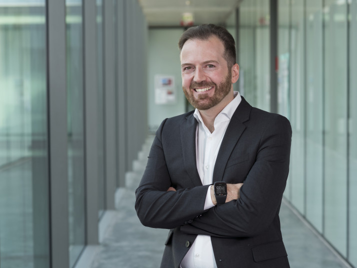 Ritratto di un uomo sorridente con capelli scuri corti e barba, che indossa un abito e tiene le braccia incrociate, in un corridoio moderno con pareti in vetro.