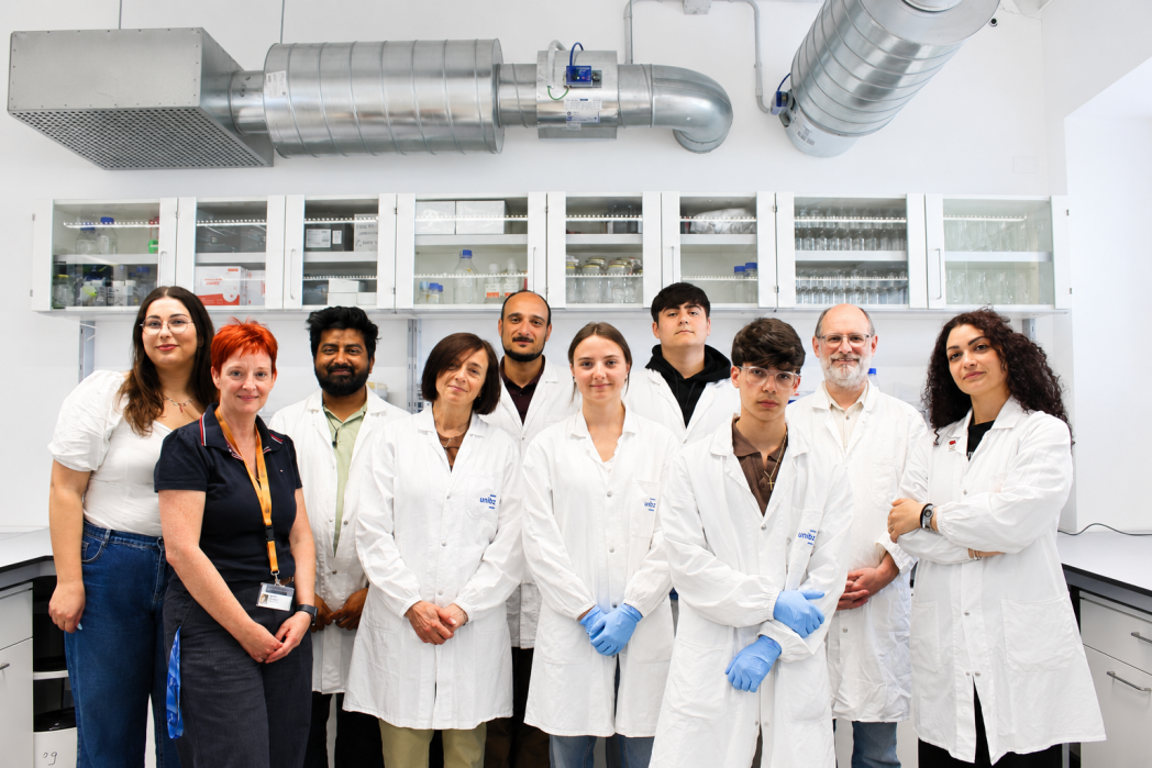 Gruppo di nove persone in camice bianco posa in un laboratorio davanti a scaffali con attrezzature e vetreria da laboratorio, guardando verso la fotocamera. 