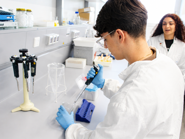 Persona con camice bianco e occhiali di protezione utilizza una pipetta in un laboratorio; sullo sfondo. pipettiert eine Flüssigkeit in einem Labor; im Hintergrund steht eine weitere Person in Laborkleidung.