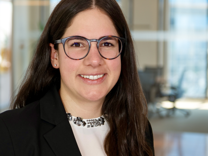 Porträt einer lächelnden Frau mit langen dunklen Haaren und runder Brille, in schwarzem Blazer und heller Bluse, vor einem unscharfen Bürohintergrund.