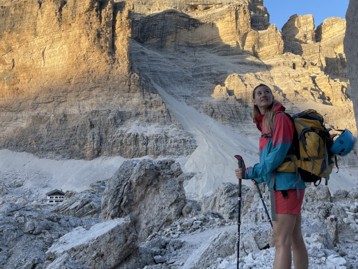 Person mit Rucksack und Wanderstöcken steht in einer felsigen Hochgebirgslandschaft vor einer steilen Felswand und einem Gletscherrest unter blauem Himmel.