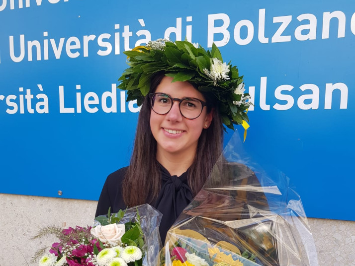 Lächelnde Absolventin mit Lorbeerkranz und Brille hält zwei Blumensträuße vor einem Schild der Universität Bozen.  