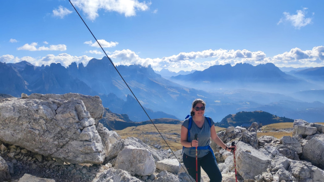 Frau mit Sonnenbrille und Rucksack wandert mit Trekkingstöcken zwischen Felsen auf einem Berg, im Hintergrund weite Berglandschaft unter blauem Himmel. Foto: Privat