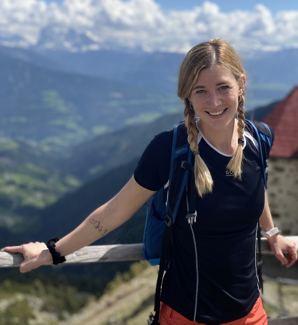 Lächelnde Person mit Zopf, Rucksack und Sportkleidung lehnt an einem Holzgeländer auf einem Aussichtspunkt, mit grünen Tälern und Bergen im Hintergrund.