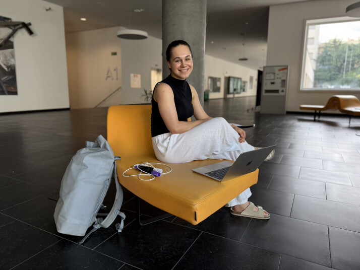 Eine Person sitzt mit einem Laptop auf einem gelben Sitzmöbel im Gebäude der Freien Universität Bozen. Ein Springseil und ein Rucksack liegen daneben.