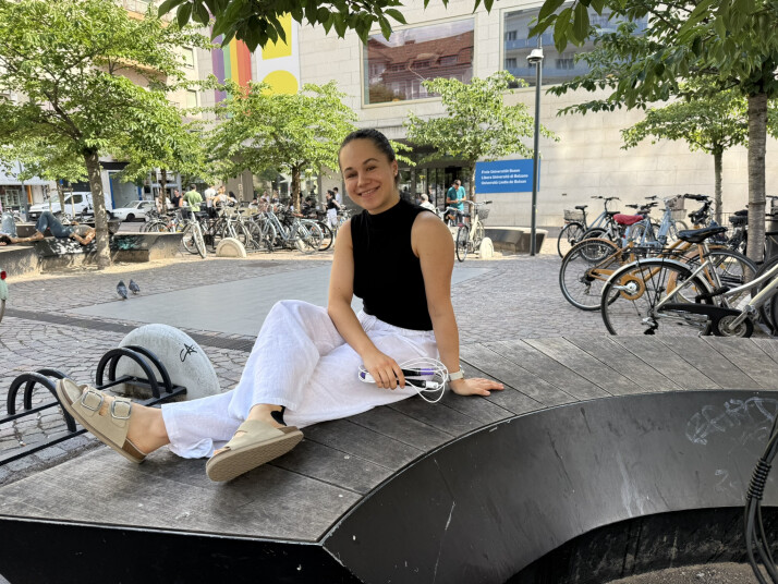 Eine Person sitzt mit einem Springseil auf einer Holzbank im Freien vor dem Gebäude der Freien Universität Bozen und lächelt in die Kamera.