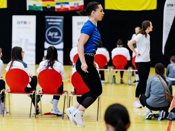 Eine Person springt in einer Sporthalle Seil, während im Hintergrund Teilnehmende und Betreuende auf einem Wettkampfgelände sitzen und stehen.