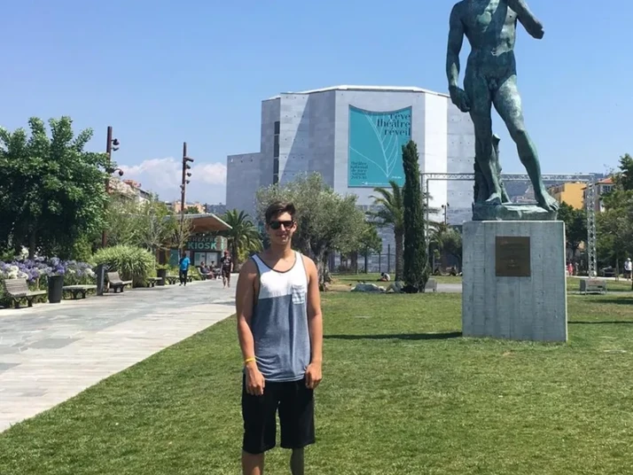 Person steht auf einer Wiese in einem Park neben einer großen Bronzestatue eines nackten Mannes auf einem Sockel; im Hintergrund ein moderner Museumsbau und ein Weg mit Bäumen.