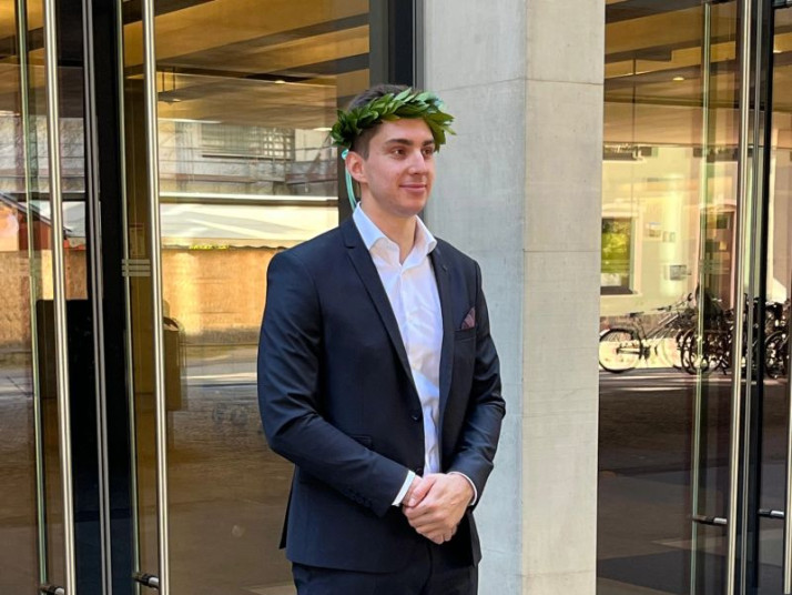 Person in dunklem Anzug mit weißem Hemd und Lorbeerkranz auf dem Kopf steht vor einem modernen Gebäude mit Glasfassade, die Hände vor dem Körper gefaltet.