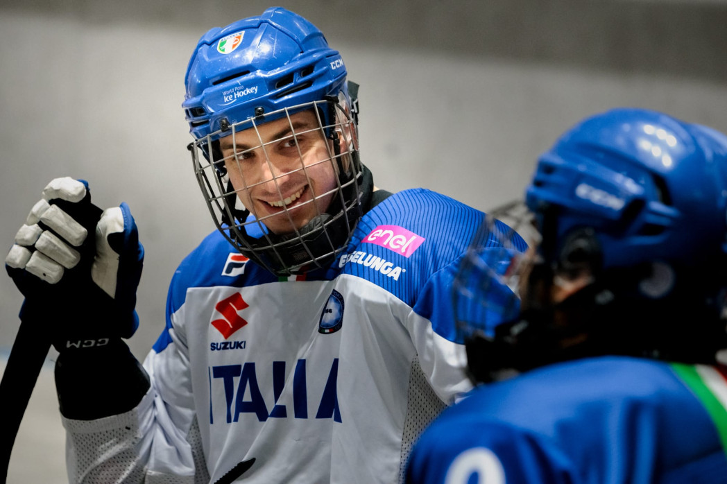 Eishockeyspieler der italienischen Nationalmannschaft mit blauem Helm und weiß-blauem Trikot mit der Aufschrift „Italia“, lächelt und hebt eine behandschuhte Hand, während er mit einem Teamkollegen spricht..