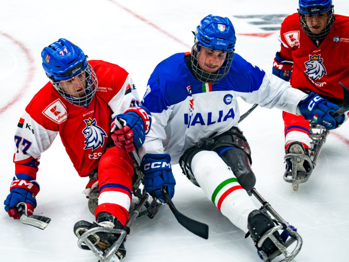 Para-Eishockeyspieler der italienischen Mannschaft sitzt auf einem Schlitten auf dem Eis und bewegt sich mit zwei kurzen Schlägern in Richtung eines Pucks; im Hintergrund ein weiterer Spieler.