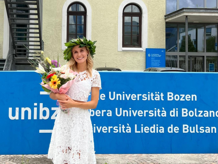 Lächelnde Frau mit Blumenstrauß und Lorbeerkranz steht vor einem Universitätsgebäude mit blauem Schild.