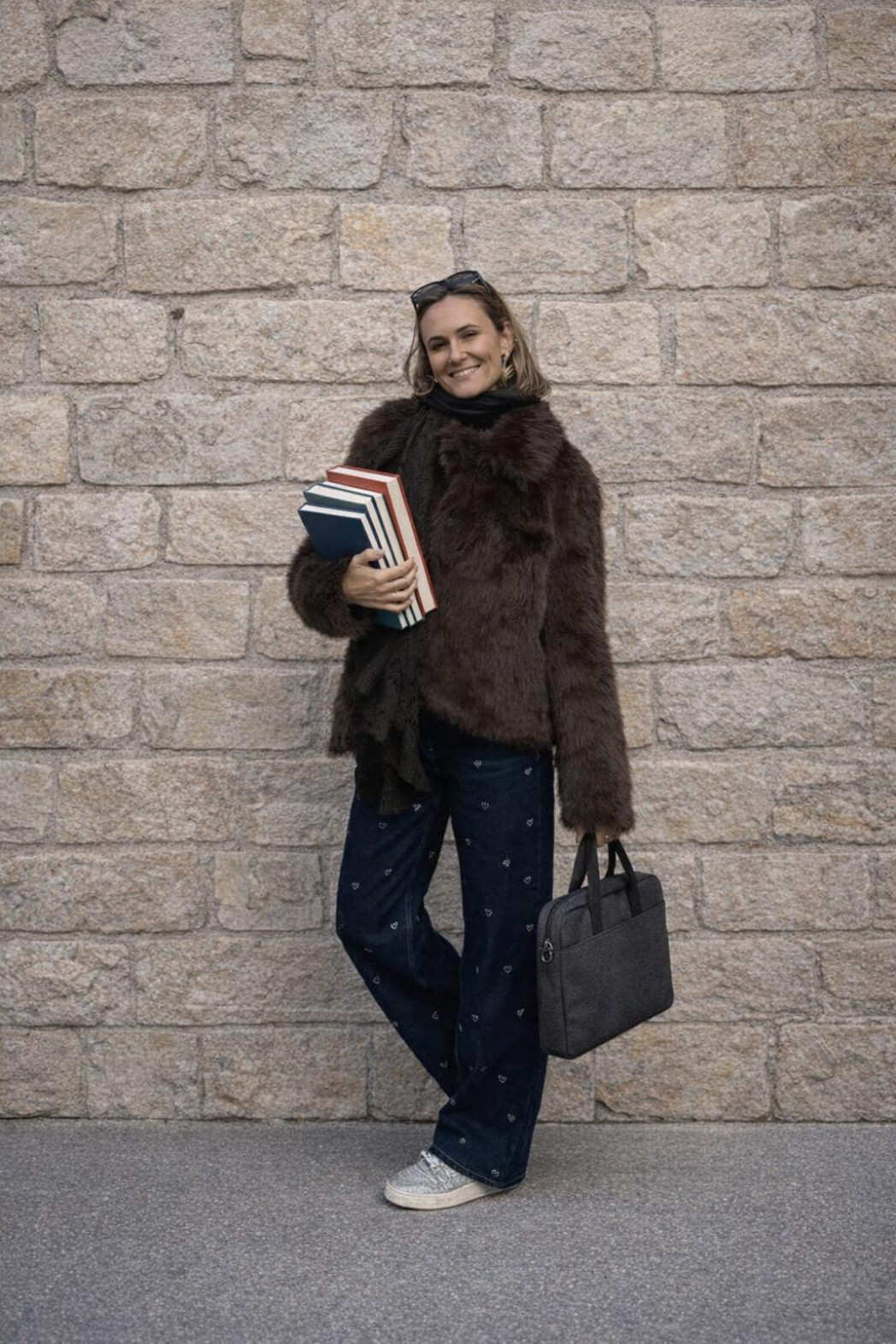 Lächelnde Frau steht vor einer Steinwand, hält mehrere Bücher im Arm und eine Tasche in der Hand.