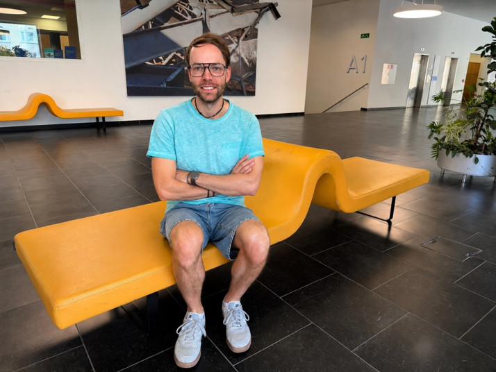 Eine Person sitzt auf einer gelben Sitzbank im Foyer der Freien Universität Bozen und blickt lächelnd in die Kamera.