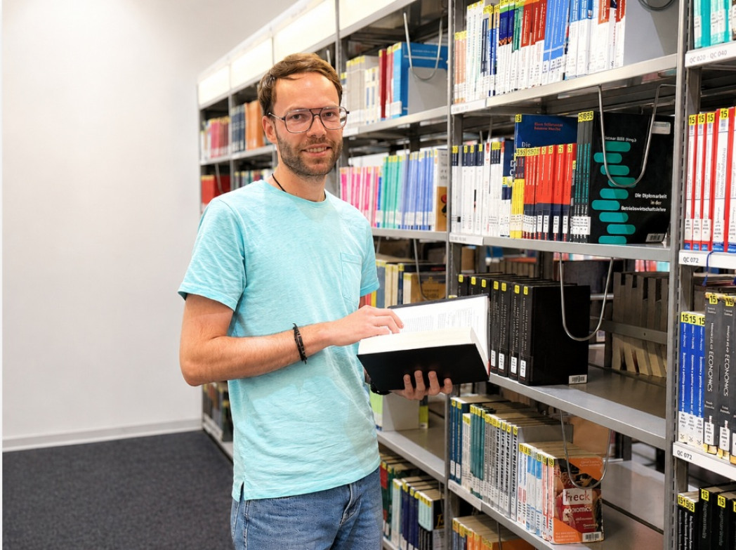 Eine Person steht in einer Bibliothek vor einem Bücherregal, hält ein aufgeschlagenes Buch in der Hand und blickt lächelnd in die Kamera.