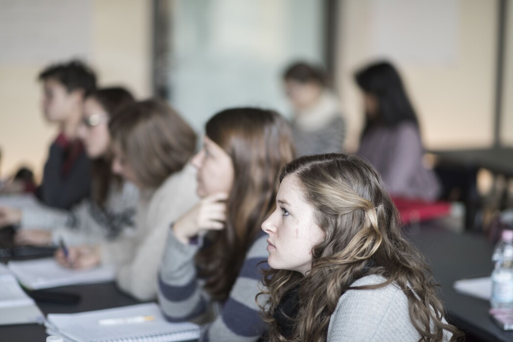 Studenti seduti attorno a un tavolo in un’aula seguono con attenzione una lezione; in primo piano si vede una persona di profilo.