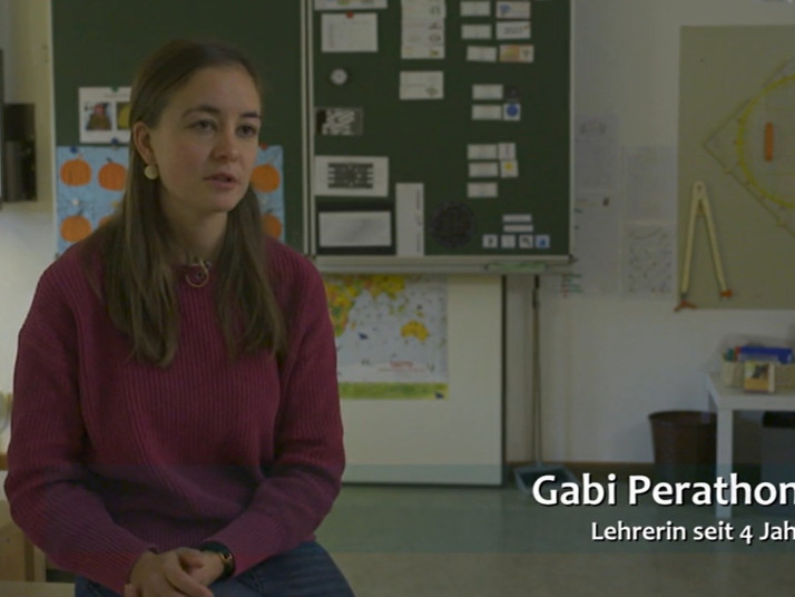 Junge Lehrerin sitzt in einem Klassenzimmer vor einer Tafel mit Unterrichtsmaterialien und spricht in die Kamera.