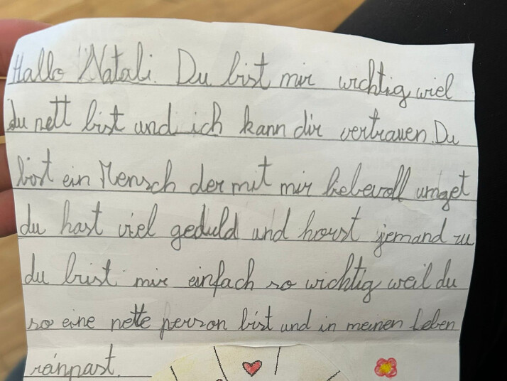 Handgeschriebener Brief auf linierten Papier mit liebevoller Nachricht an „Natali“. Unten ist eine bunte Kinderzeichnung einer Person mit ausgebreiteten Armen, umgeben von Herzen, Blumen und Strahlen.