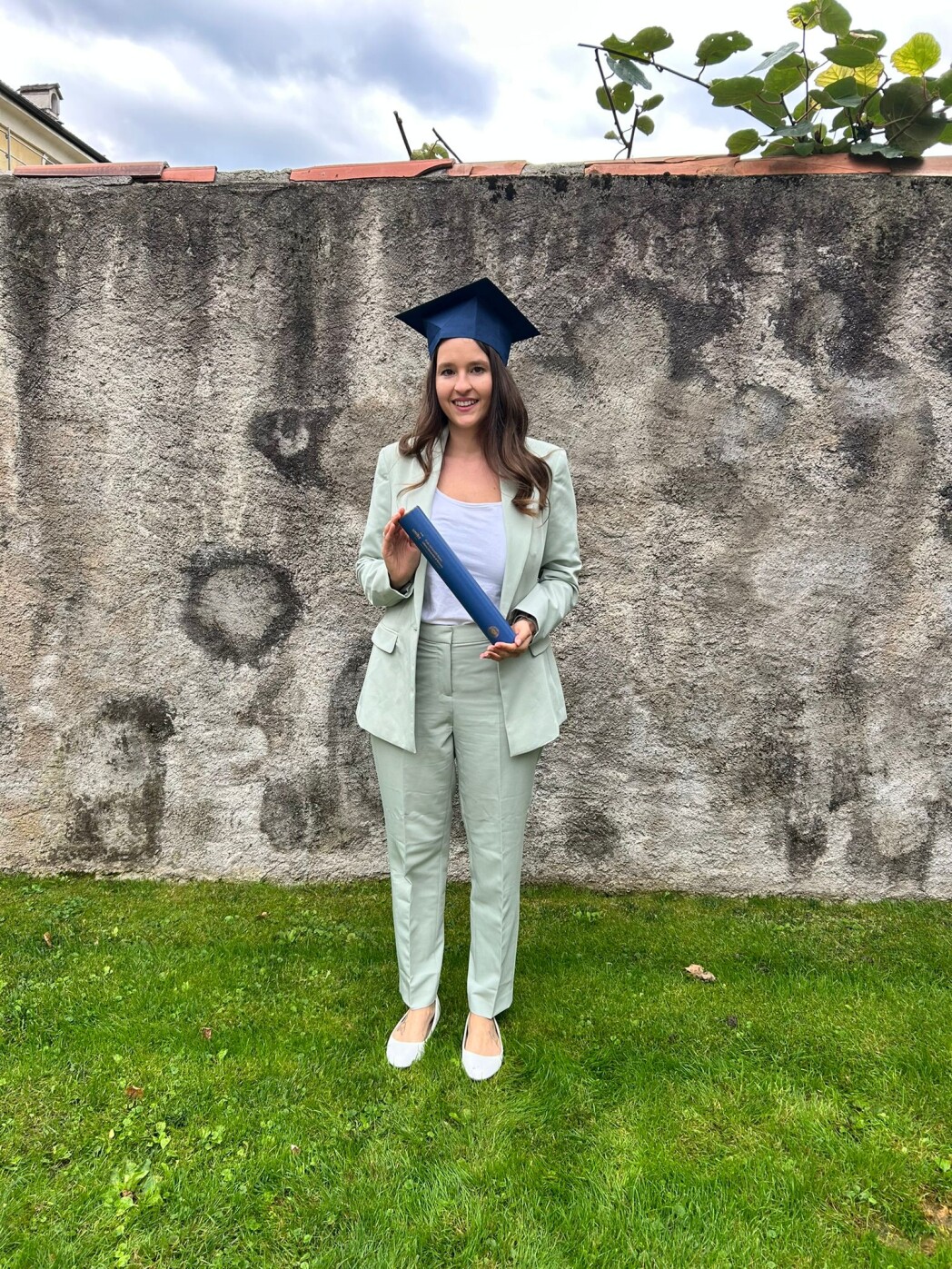 Junge Frau in hellgrünem Hosenanzug und Abschlusskappe steht auf einer Wiese vor einer grauen Mauer und hält eine blaue Diplomrolle in der Hand.