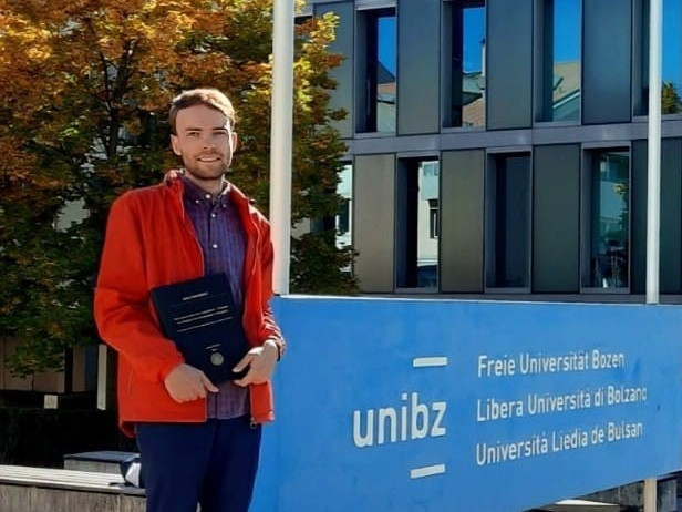 Eine Person steht vor dem Eingangsbereich der Freien Universität Bozen neben einem unibz-Schild und hält eine Urkunde oder Mappe in den Händen.