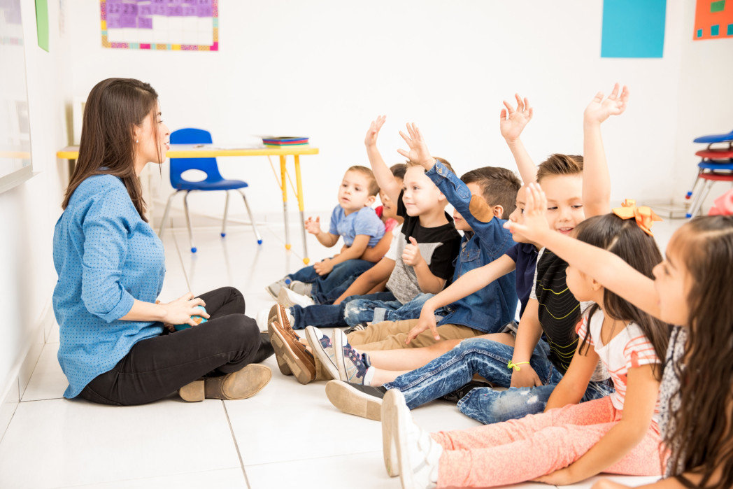 Eine Frau sitzt auf dem Boden vor Kindern. Sie halten eine Hand in die Höhe.