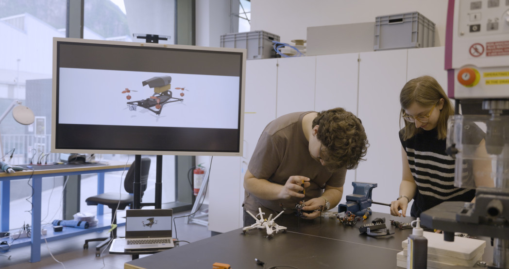 Due studenti lavorano in laboratorio su componenti elettronici; sul tavolo ci sono strumenti e parti di un piccolo drone, mentre un monitor sullo sfondo mostra un modello 3D di un drone.