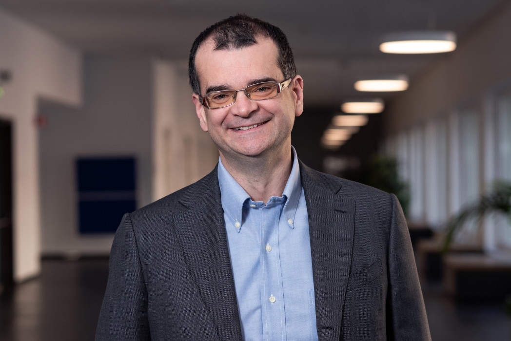 Ritratto professionale di uomo sorridente con occhiali in blazer scuro e camicia azzurra in ufficio moderno.