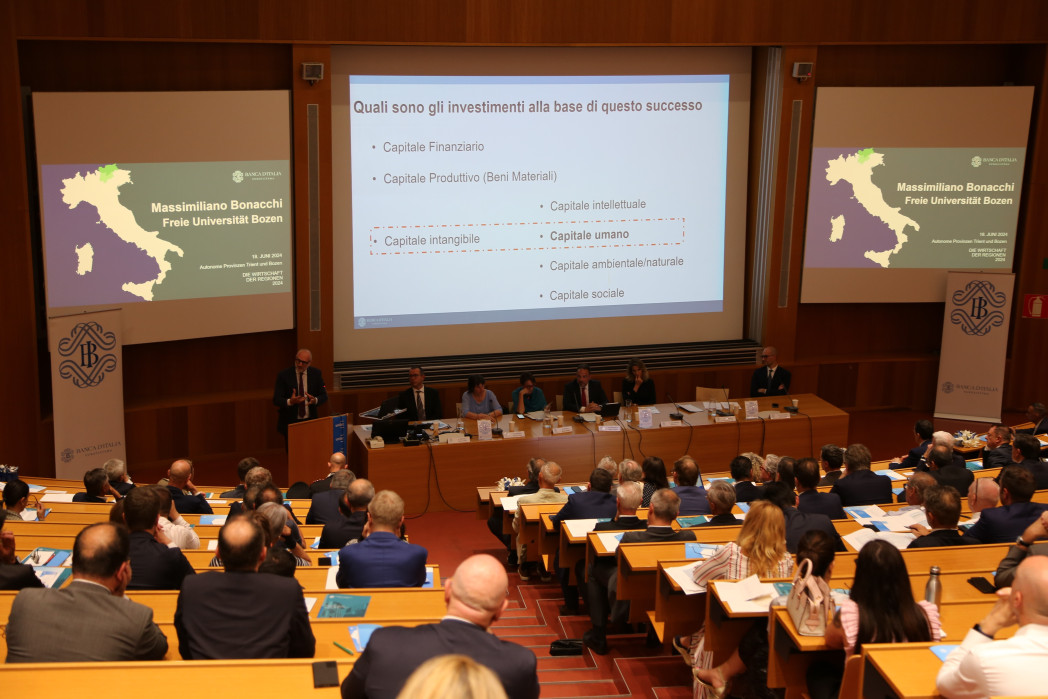 Una sala conferenze con molte persone. tre diapositive proiettate sulla parete. Una persona parla su un podio. 