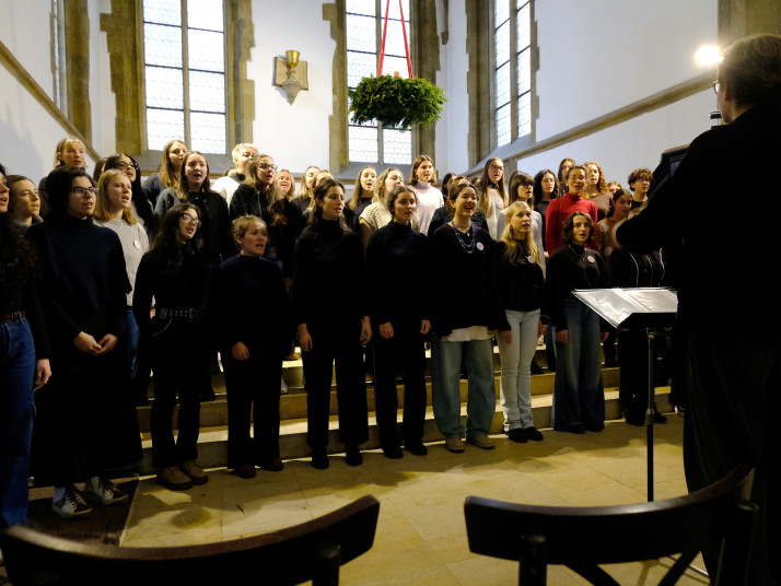 A choir stands at the front of a church, singing together under tall arched windows.