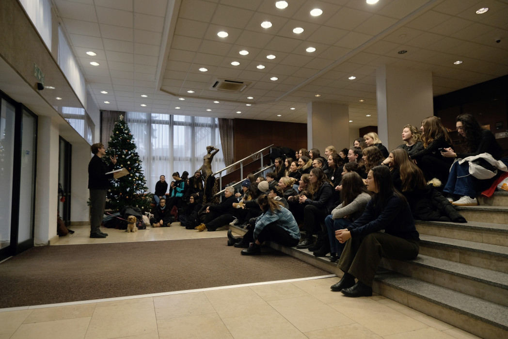 A group of people sits on indoor steps, listening to a speaker near a decorated Christmas tree.
