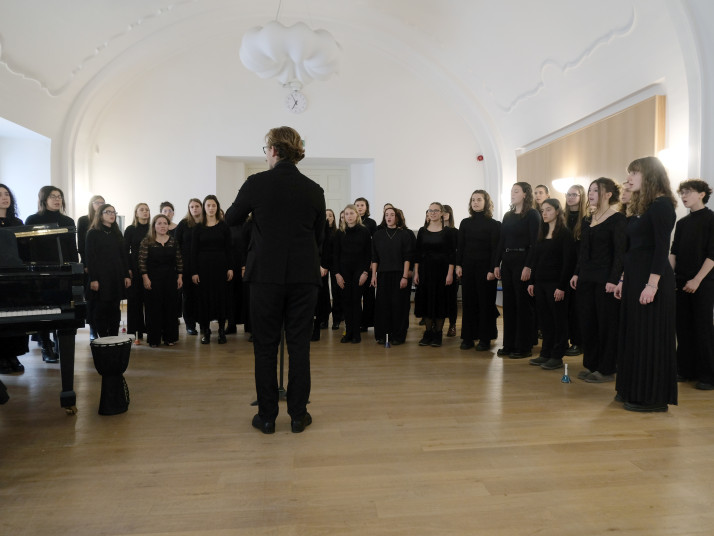 A university choir dressed in black performs in a bright concert hall, led by a conductor standing in front of the singers.