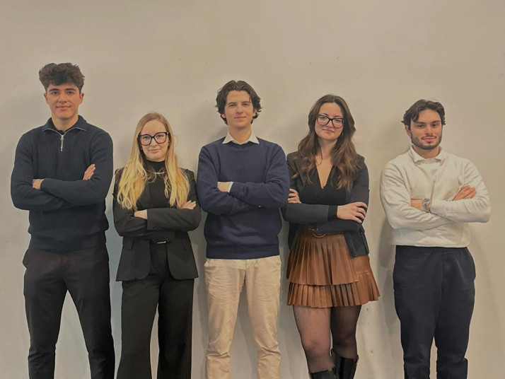 Five young adults stand side by side with arms crossed in front of a plain, light-colored indoor wall.