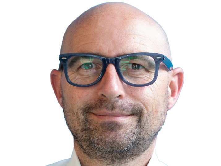 Ritratto di un uomo sorridente, calvo, con occhiali e camicia bianca su sfondo chiaro.