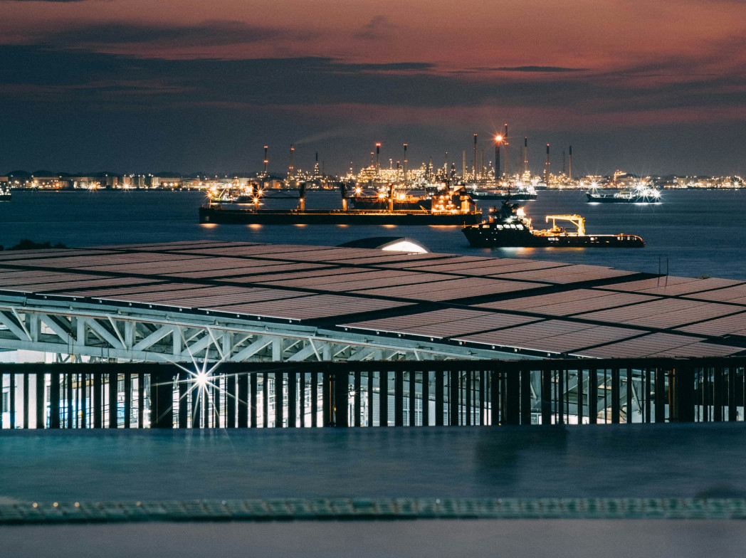Porto industriale di notte con navi illuminate e impianti di raffineria sullo sfondo; in primo piano il tetto di un edificio moderno.