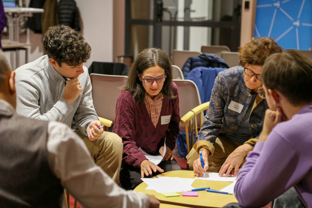 Un gruppo di persone sedute attorno a un tavolo discute e lavora concentratamente con appunti e cartoncini colorati.