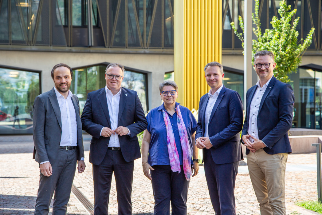 Eine Gruppe von fünf Personen steht im Freien vor einem modernen Gebäude, sie lächeln in die Kamera.