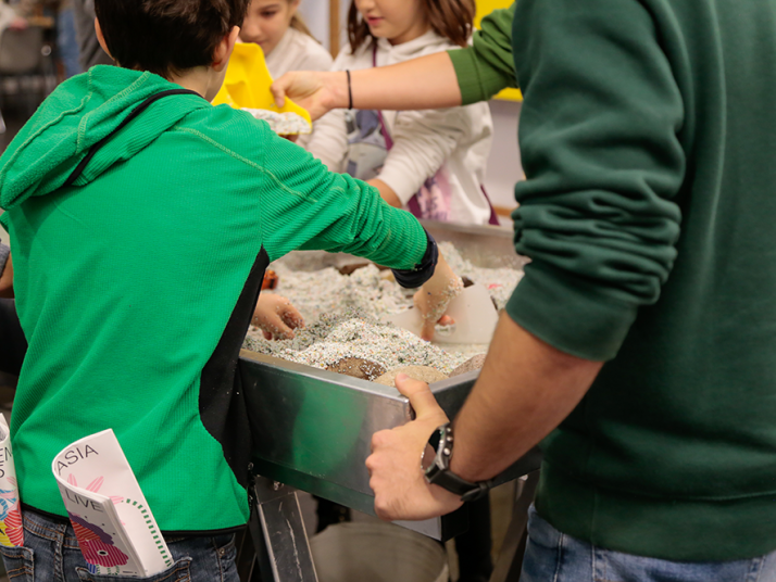 Kinder in grünen Sweatshirts nehmen an praktischer Sinnesaktivität mit Sand oder ähnlichem Material in einem Behälter teil.