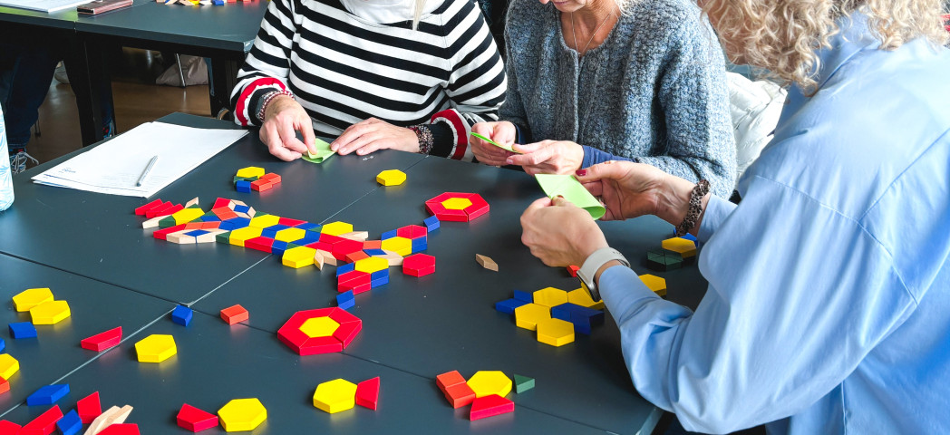 Drei Personen sitzen an einem Tisch und legen bunte geometrische Spielsteine aus Holz aus.