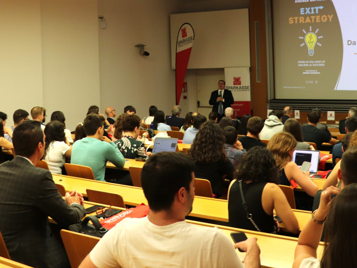 Un uomo in piedi parla, mentre le persone sedute nell'aula lo ascoltano.
