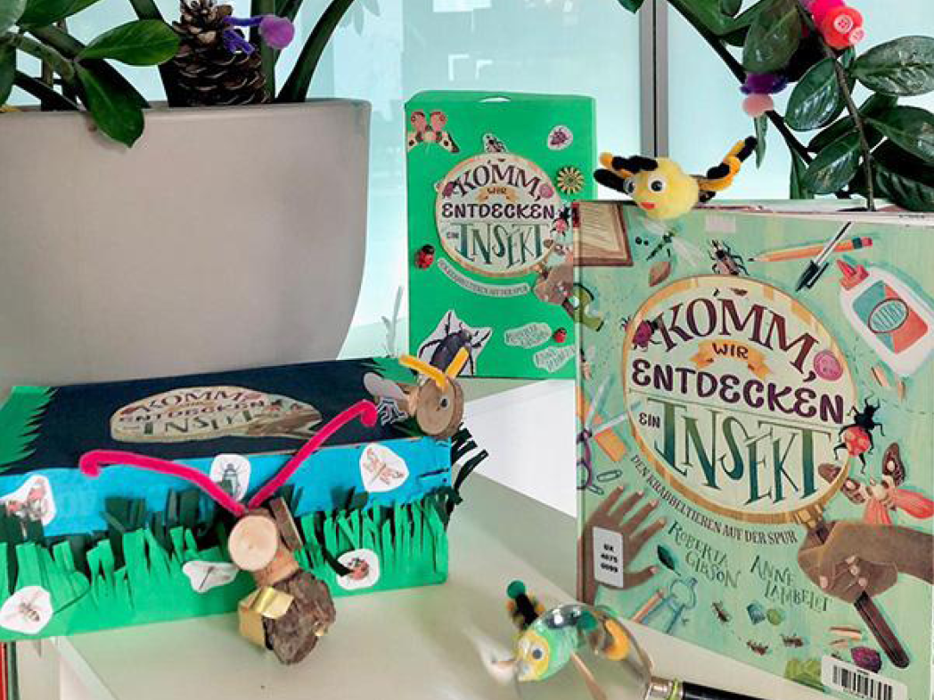  Tisch, auf dem ein Kinderbuch zum Thema Insekten, eine kreativ gestaltete Bücherkiste, eine Zimmerpflanze mit gebastelten Insekten sowie eine Lupe zu sehen sind.