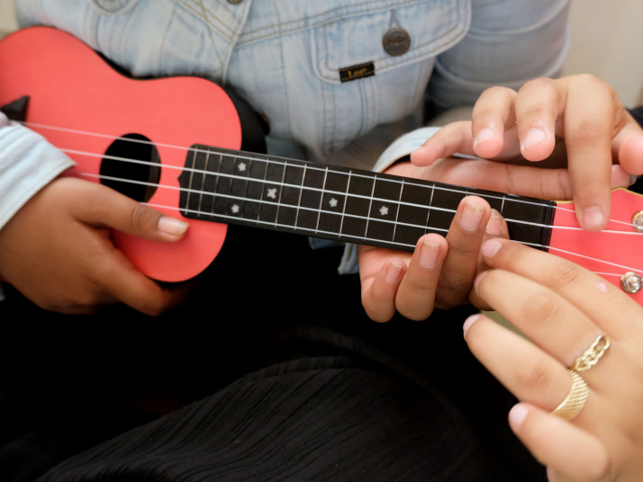 Vier Kinderhände auf einer Ukulele. Eine Kind zeigt dem anderen die Griffe.