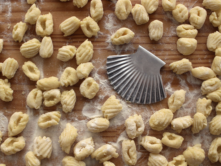 Gnocchibrett mit vielen grillten Gnocchi. In der Mitte liegt ein Küchenwerkzeug aus Edelstahl in Form einer Jakobsmuschel mit dem man die Gnocchi schneidet und die Rillen formt.