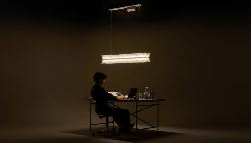 Eine Person sitzt an einem Schreibtisch in der Mitte eines relativ dunklen Raumes - darüber hängt eine Hängeleuchte, die nach oben und untern leuchtet