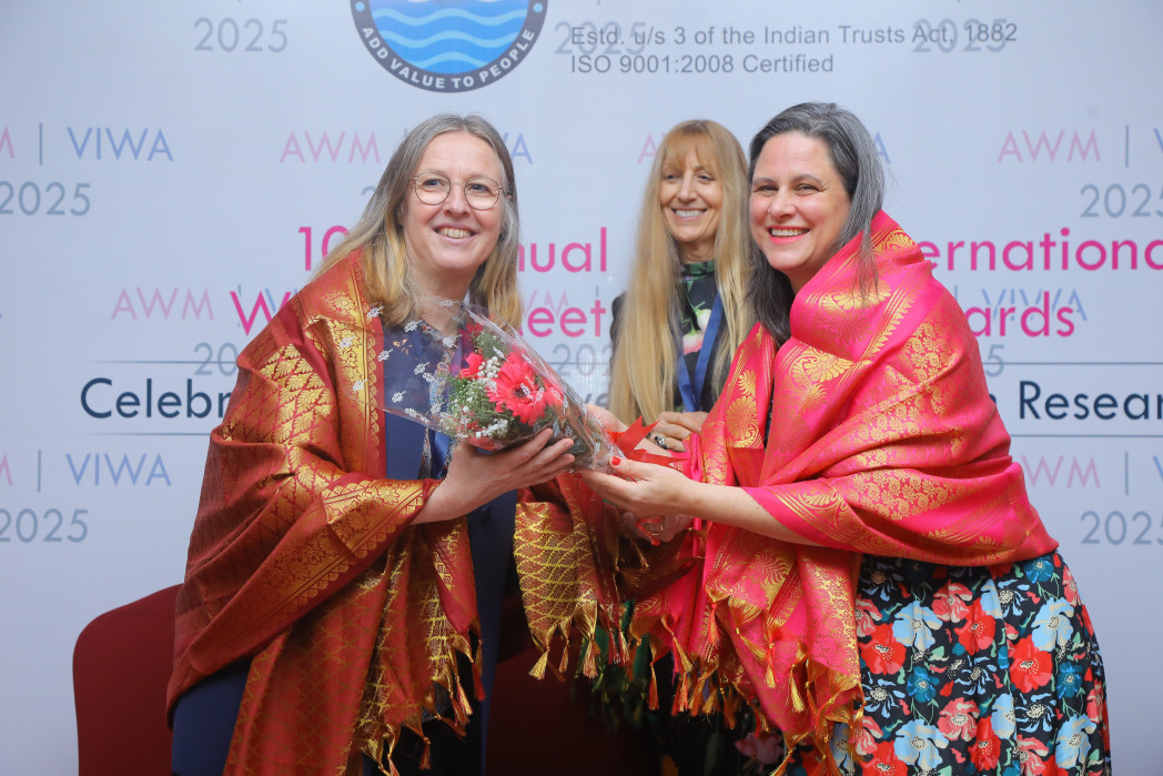 Drei Frauen in bunten Kleidern, die Preisträgerin bekommt einen Blumenstrauß überreicht.