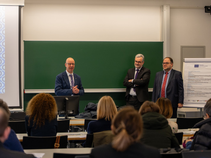 Drei Männer stehen vorne in einem Hörsaal vor einer grünen Tafel und sprechen zu einem sitzenden Publikum.
