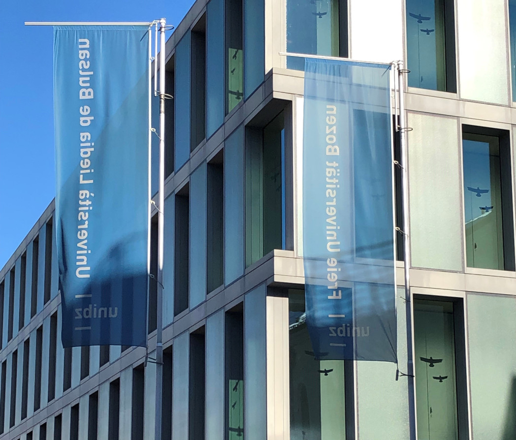 Zwei blaue Fahnen mit der Aufschrift „Freie Universität Bozen“ vor der modernen Glasfassade eines Universitätsgebäudes.