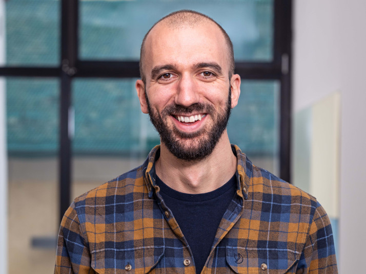 Ritratto di un uomo sorridente con capelli corti e barba, che indossa una camicia a quadri, davanti a un edificio moderno con facciata in vetro.