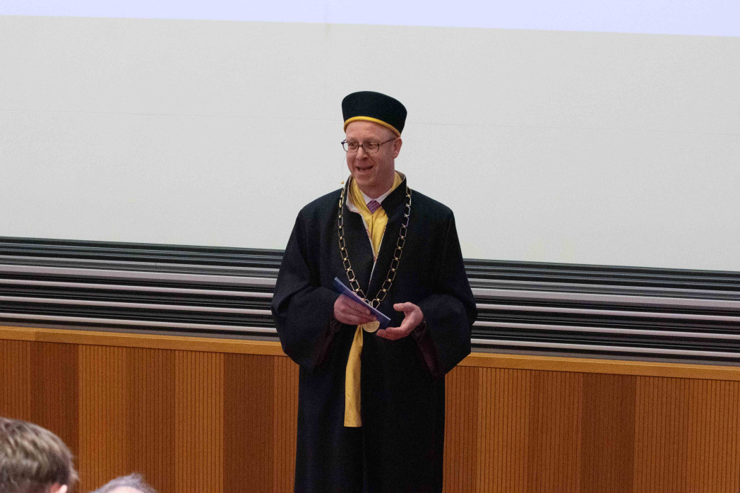 Una persona in toga accademica con collare cerimoniale e copricapo è in piedi in un’aula e tiene degli appunti in mano. Sullo sfondo si vedono pannelli in legno e uno schermo di proiezione chiaro.