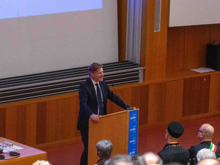 Un uomo in giacca parla da un podio in un’aula universitaria. Davanti a lui c’è il pubblico; l’ambiente è rivestito in legno, con uno schermo di proiezione sullo sfondo.