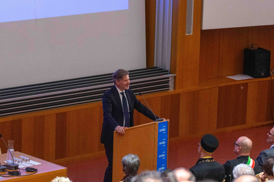 Un uomo in giacca parla da un podio in un’aula universitaria. Davanti a lui c’è il pubblico; l’ambiente è rivestito in legno, con uno schermo di proiezione sullo sfondo.
